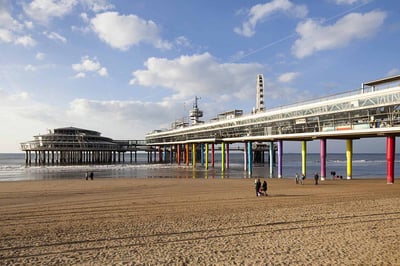 Scheveningen Pier on the beach in The Hague, Netherlands, featuring colorful pillars, a Ferris wheel, and a bungee jump tower stretching over the sea with people walking on the sandy shore.