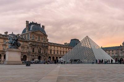 Alt text: Louvre Museum courtyard at sunset, featuring the iconic glass Louvre Pyramid and the classical Parisian architecture of the museum building under a warm sky, with visitors present.