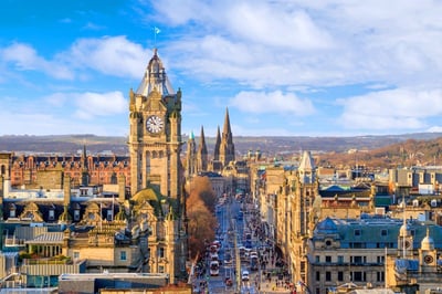Panoramic view of Edinburgh's bustling Princes Street, featuring an iconic clock tower on the left, leading towards historic spires and buildings under a bright blue sky.