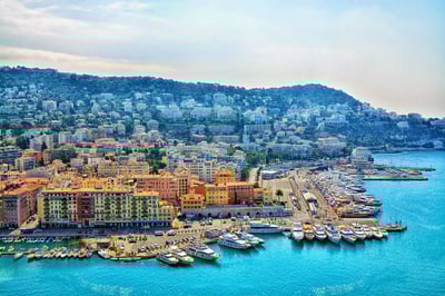 Panoramic aerial view of Port Lympia in Nice, France, showcasing turquoise waters filled with luxury yachts and boats, vibrant multi-story buildings lining the busy waterfront, and a hillside city extending into the background.
